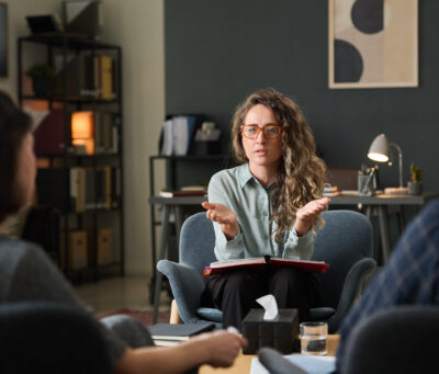 Discussing Work Strategies in Modern Office Environment Three colleagues engaging in discussion about work strategies in a modern office setting, collaborating for project advancement and sharing ideas, seated around the table, Caucasian woman talking